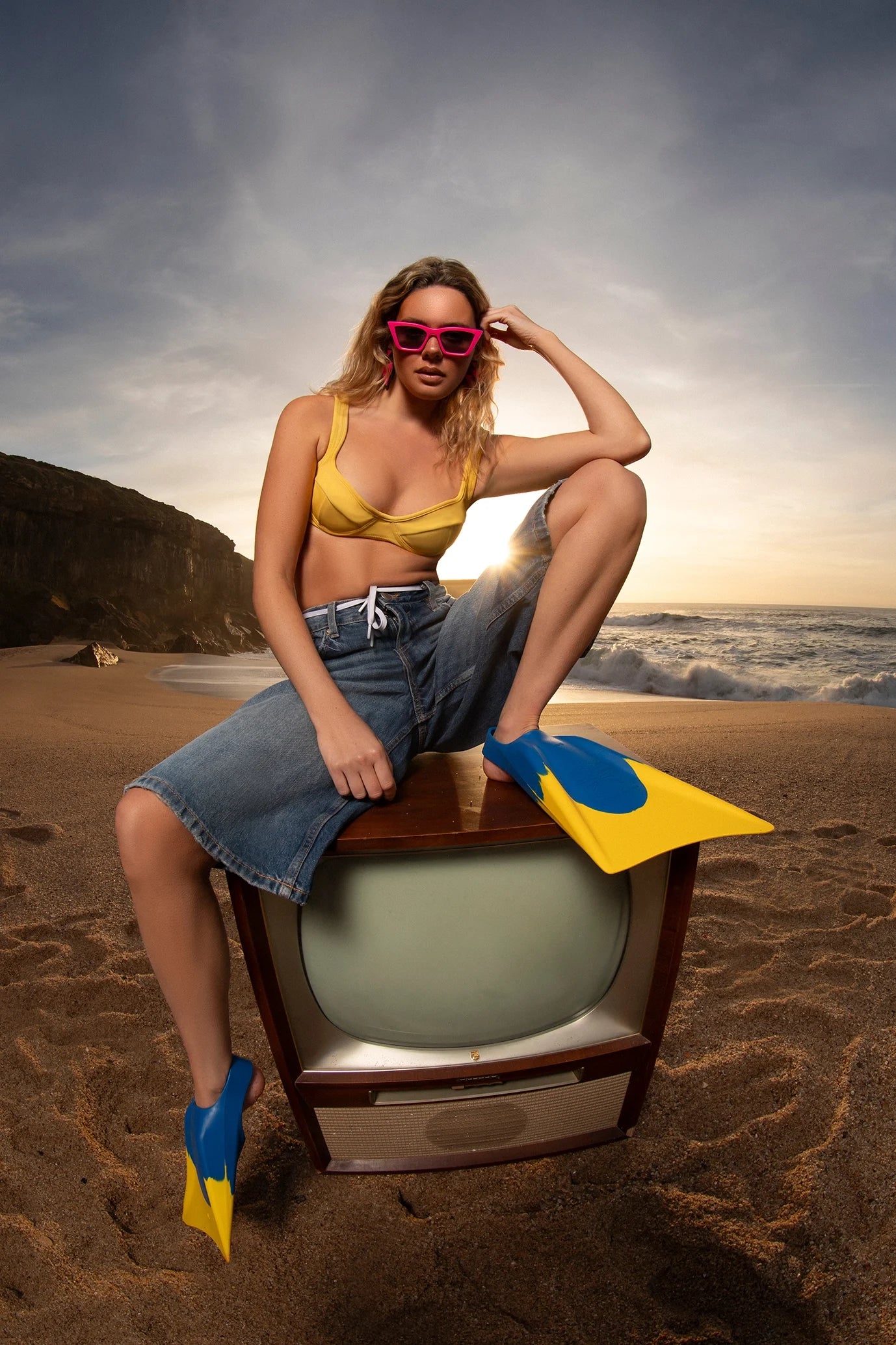 Person sitting on a vintage television set on a beach with ocean and sky in the background